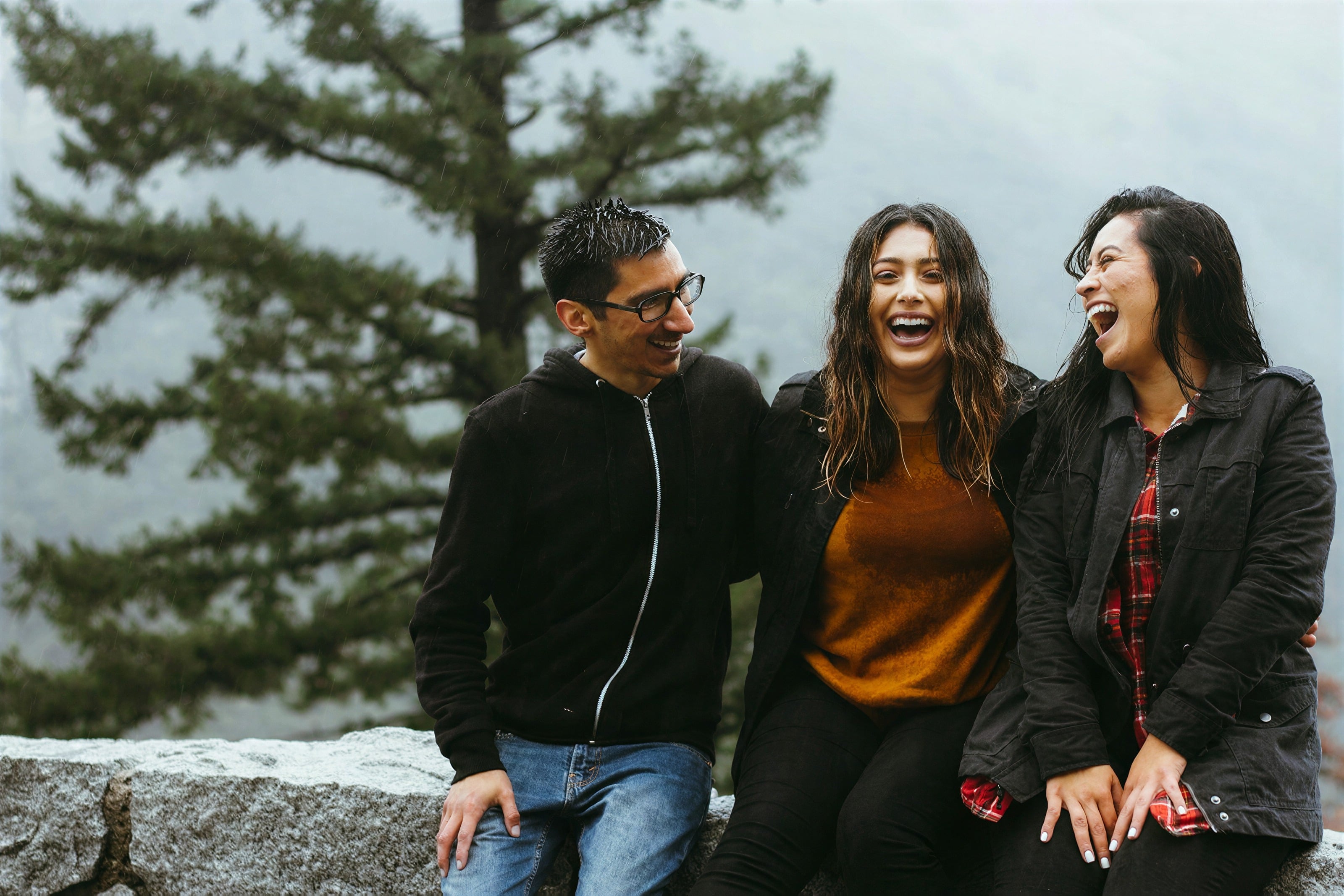 Three friends laughing together on a rock by the trees