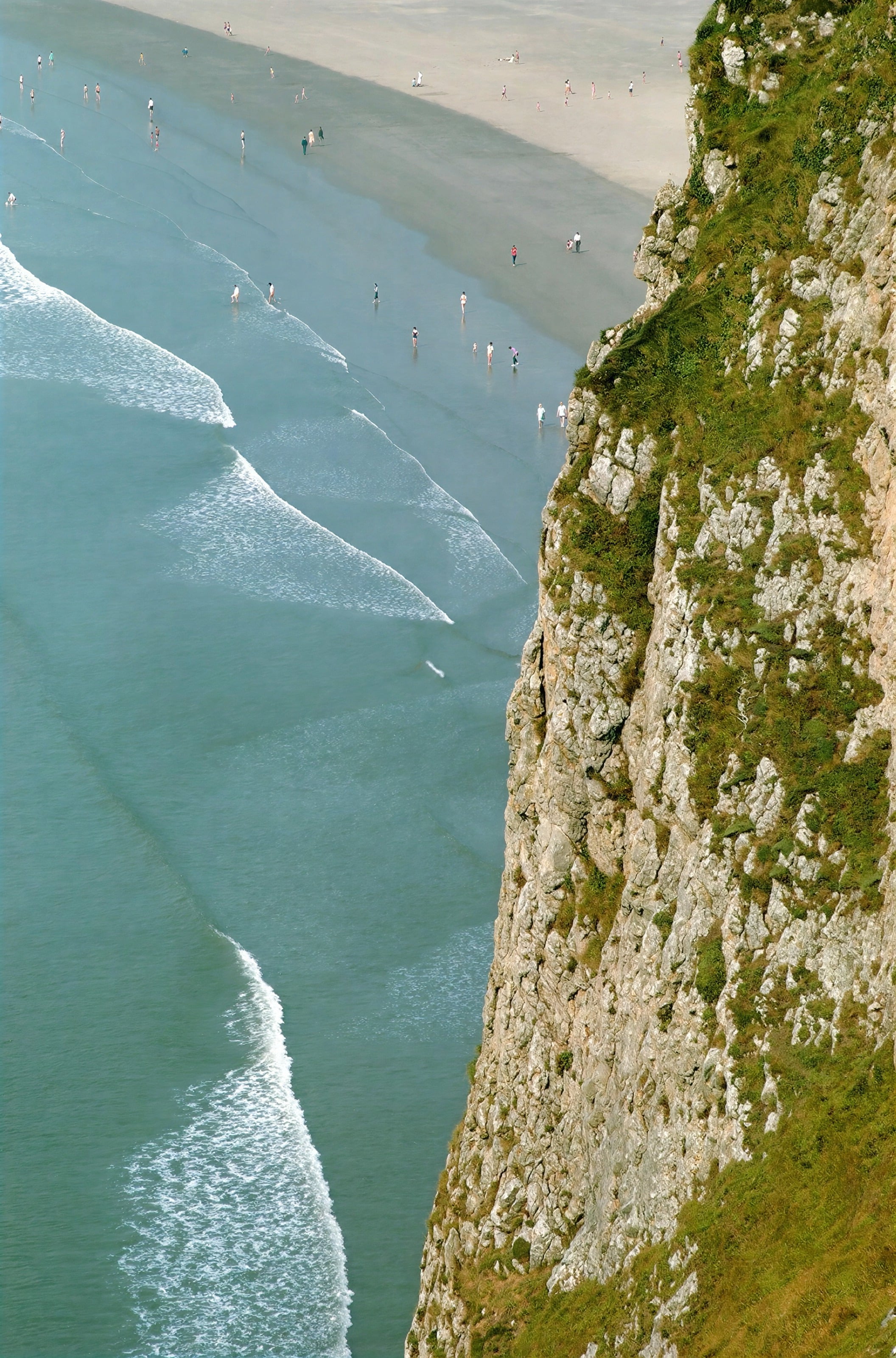 Waves rolling onto a beach below a green clifftop, seen from above